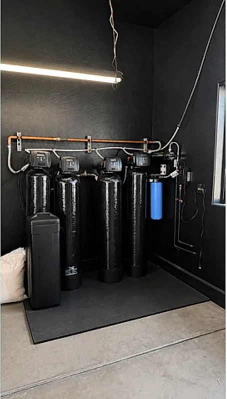 A four-tank Hydrology filtration and softening system with a brine tank and sediment filter, installed against a black wall